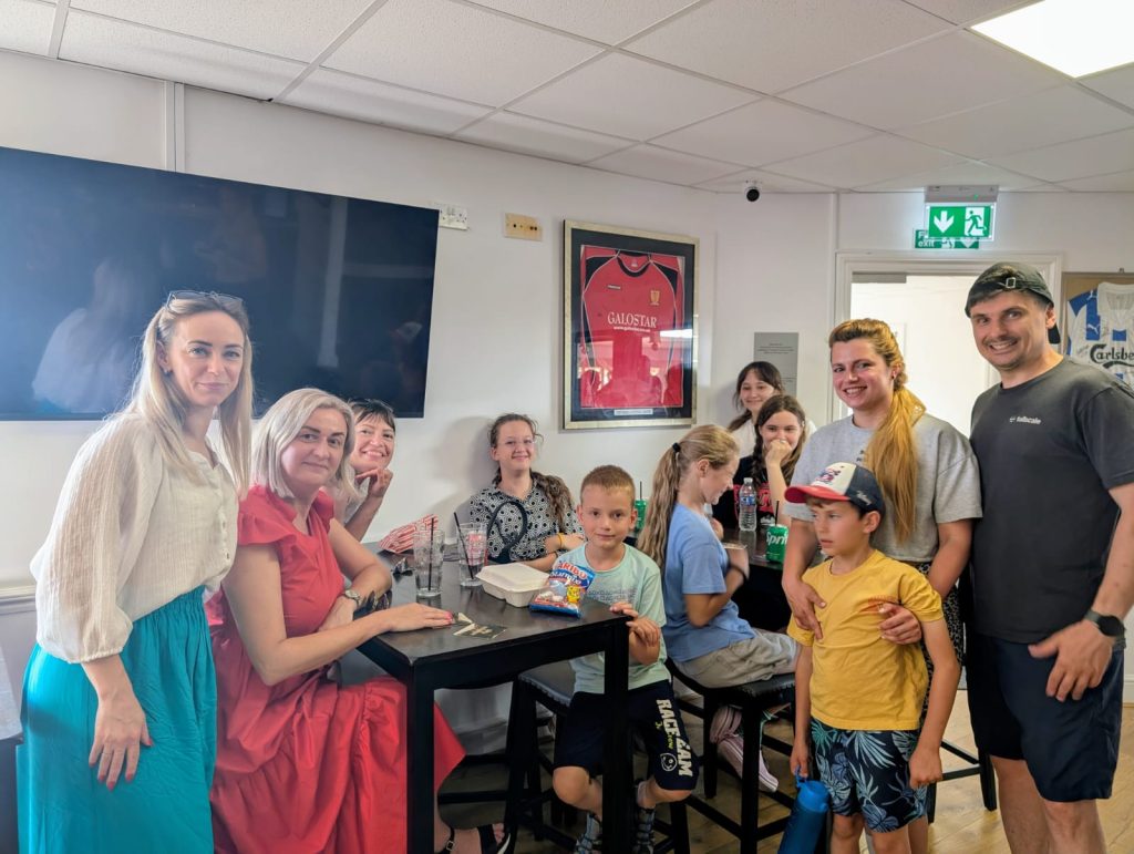 A group of people, including adults and children, gathered smiling in a room with a framed sports jersey on the wall. The scene conveys warmth and togetherness.