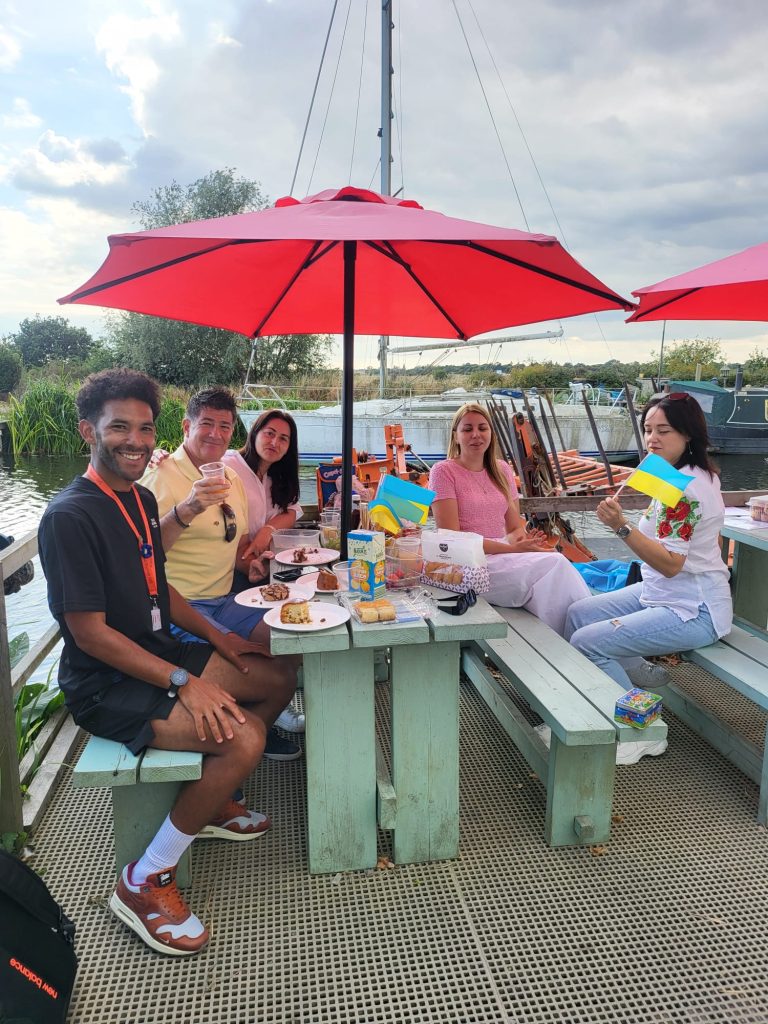 A group of five people sit around a picnic table with a red umbrella, enjoying a relaxed moment. Some hold Ukrainian flags. Plates of food are on the table, set against a scenic riverside backdrop, creating a cheerful and convivial atmosphere.