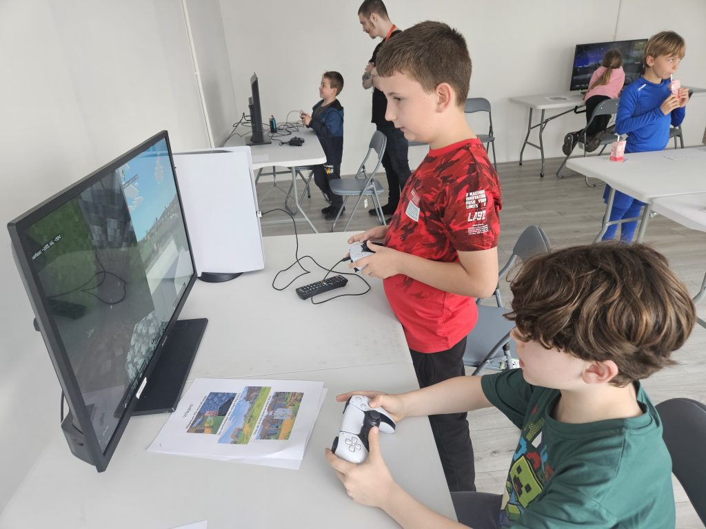 Two boys play a video game on a monitor in a bright room, holding controllers intently. Other children in the background, some using computers and drinking, create a lively, playful atmosphere.