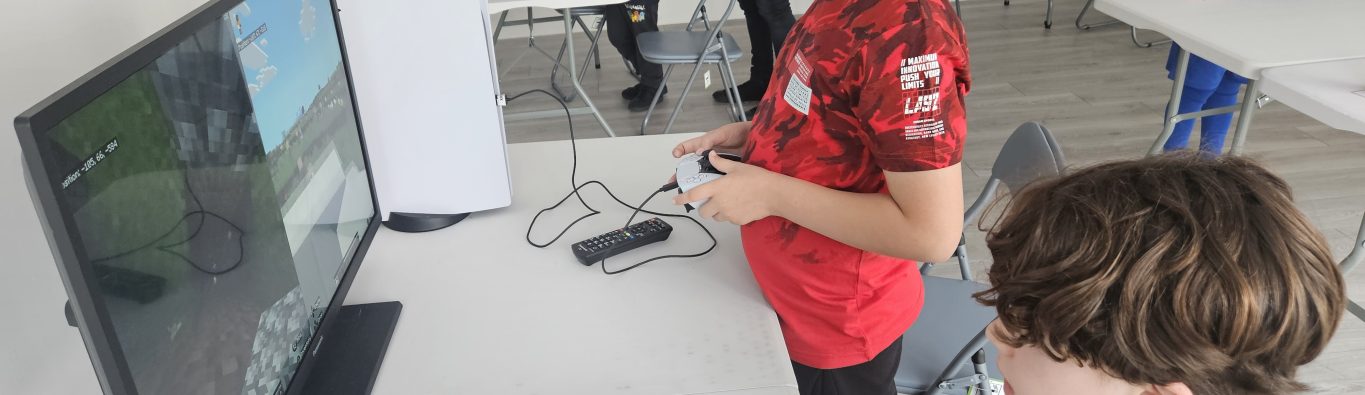 Two boys play a video game on a monitor in a bright room, holding controllers intently. Other children in the background, some using computers and drinking, create a lively, playful atmosphere.