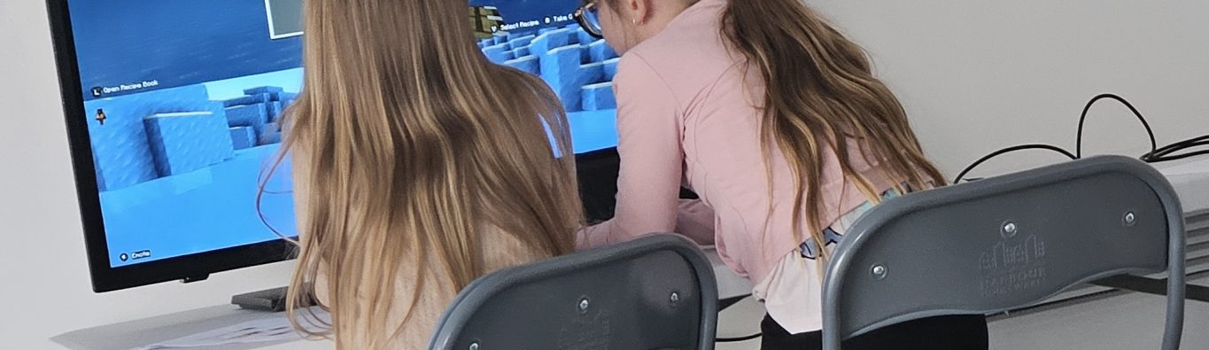 Two young girls with long hair sit and lean on chairs, focused on a computer screen displaying a block-based video game in a bright room.