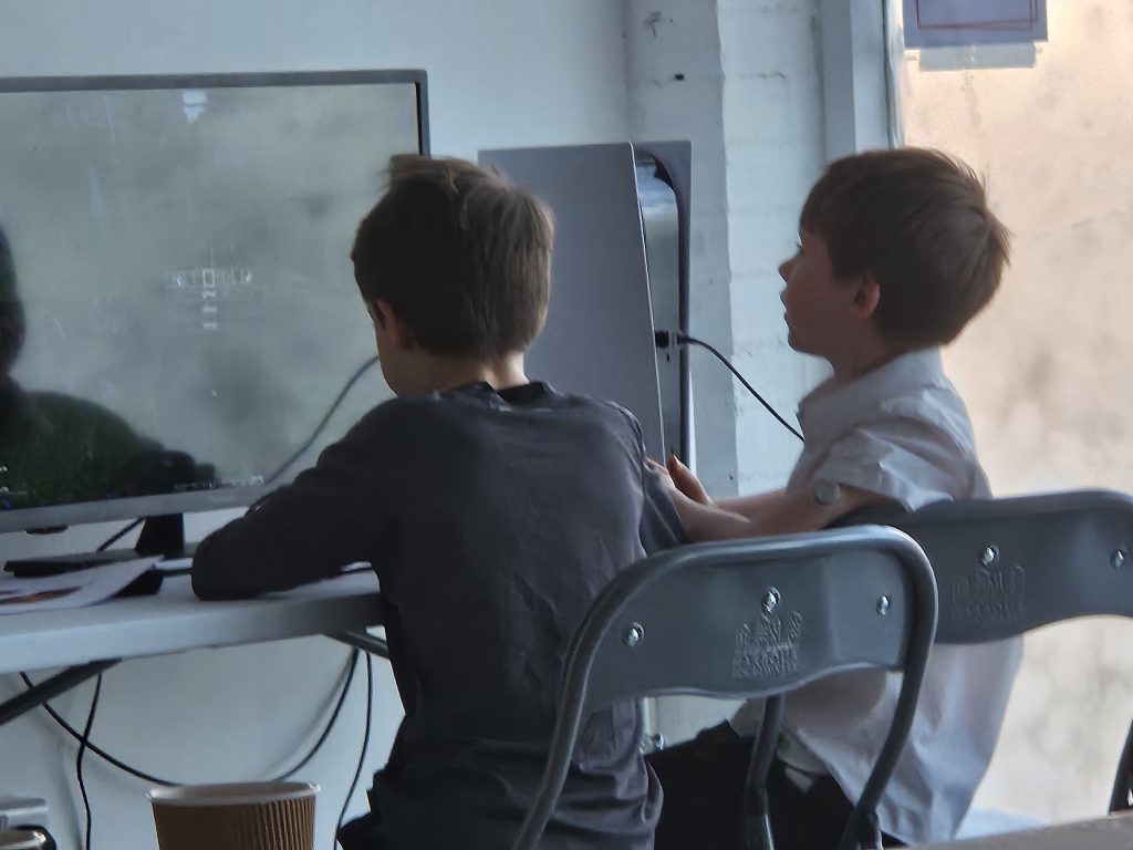 Two young boys sit on chairs at a table, focused on a video game displayed on a monitor in front of them. A relaxed, engaged atmosphere.