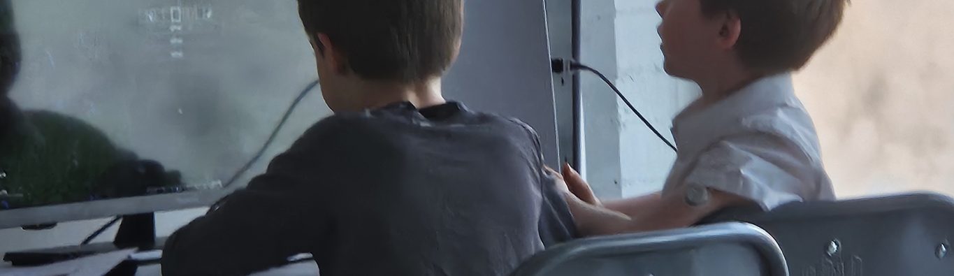 Two young boys sit on chairs at a table, focused on a video game displayed on a monitor in front of them. A relaxed, engaged atmosphere.