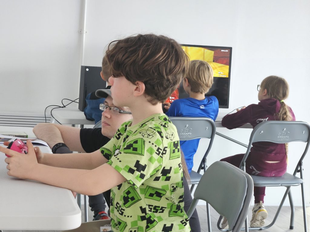 Children sit at foldable tables, intently playing video games on screens. The atmosphere is focused and engaging, with colourful clothing and game visuals.