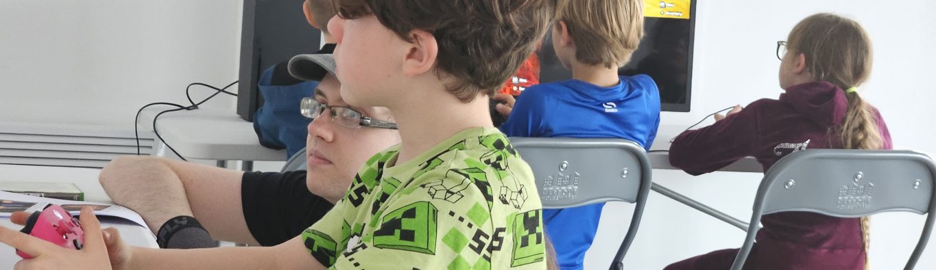 Children sit at foldable tables, intently playing video games on screens. The atmosphere is focused and engaging, with colourful clothing and game visuals.