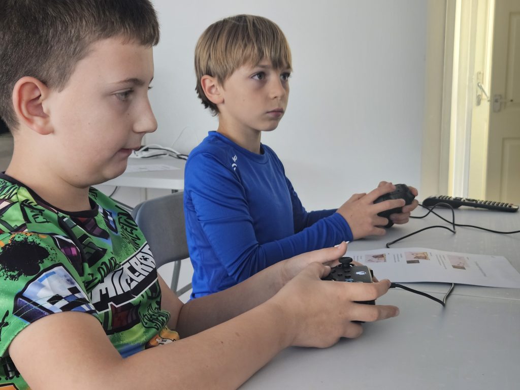 Two boys are sitting at a table focused on playing video games with controllers. They appear intent and engaged. One wears a colorful shirt; the other a blue top.