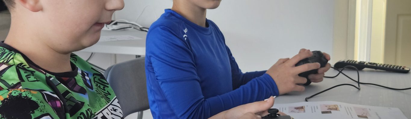 Two boys are sitting at a table focused on playing video games with controllers. They appear intent and engaged. One wears a colorful shirt; the other a blue top.