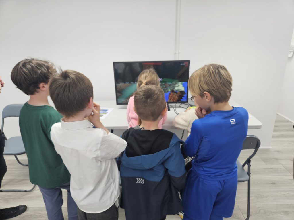 A group of children gathered around a monitor playing a colorful block-based video game. The scene conveys excitement and curiosity in a bright room.