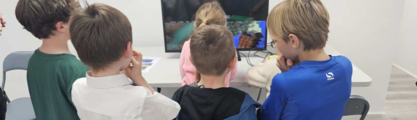 A group of children gathered around a monitor playing a colorful block-based video game. The scene conveys excitement and curiosity in a bright room.