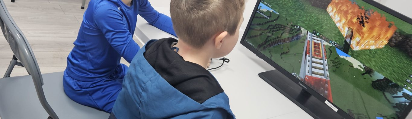 Two boys, one seated and one standing, play a video game on a monitor in a room with several computers. The mood is focused and engaging.