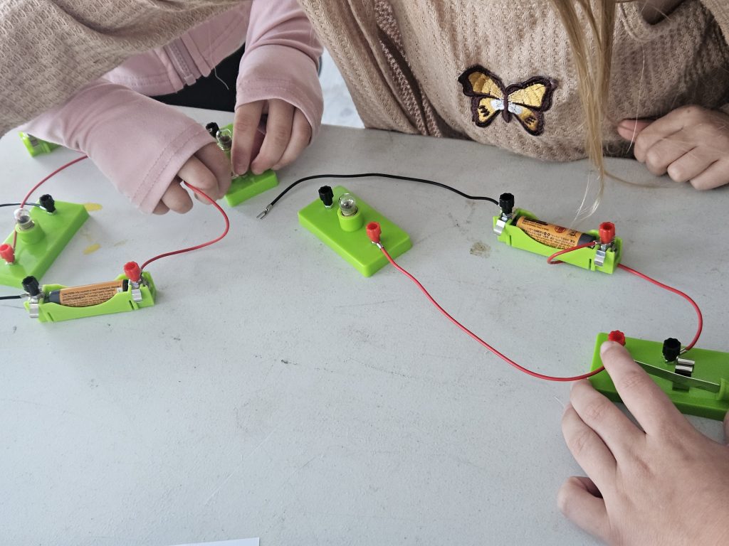 Children's hands engage with a simple circuit board setup on a table, featuring green bases, red and black wires, and switches, conveying curiosity and learning about electronics.