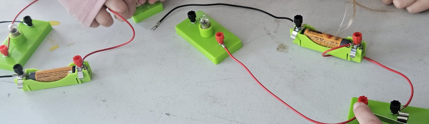 Children's hands engage with a simple circuit board setup on a table, featuring green bases, red and black wires, and switches, conveying curiosity and learning about electronics.