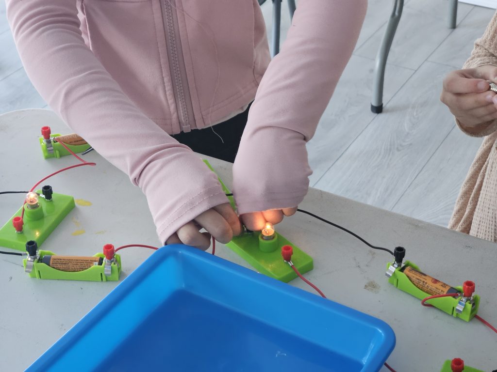 Two children in long sleeves work with a basic circuitry set on a table. The focus is on connecting wires to light up bulbs, indicating teamwork.