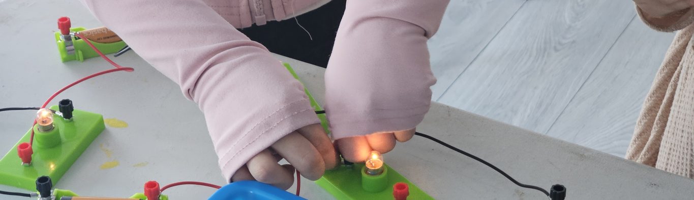 Two children in long sleeves work with a basic circuitry set on a table. The focus is on connecting wires to light up bulbs, indicating teamwork.