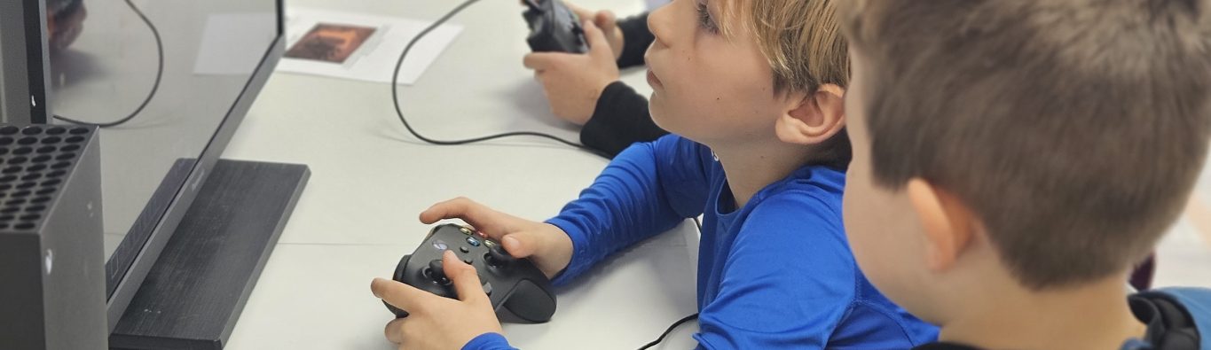 Three children sit at a table, focused and engaged, playing a video game on screens. They hold controllers, conveying concentration and excitement.