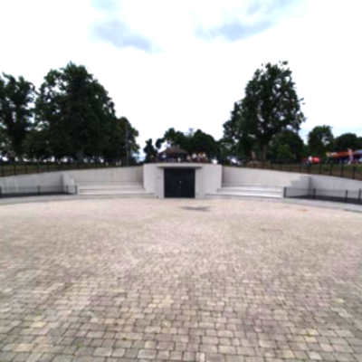 Wide cobblestone plaza leading to a modern, minimalist structure surrounded by trees. Cloudy sky adds a serene and contemplative tone.