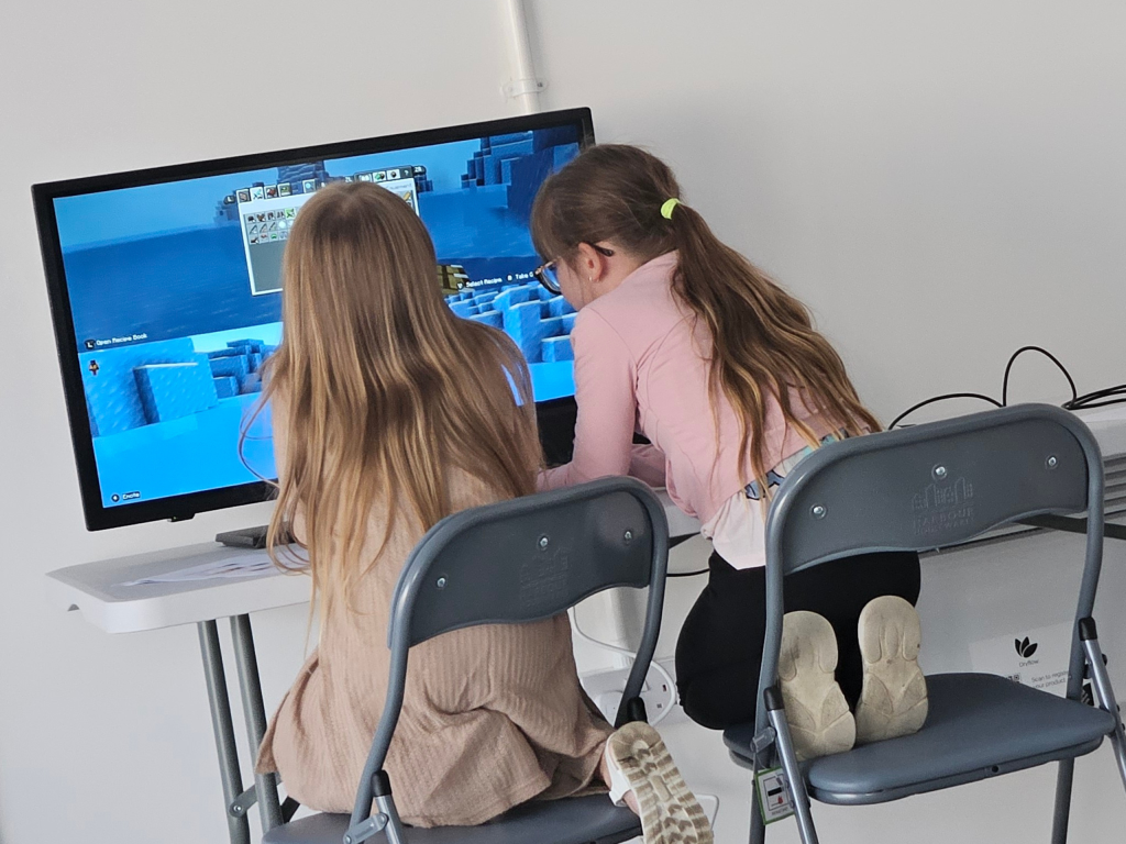 Two children in long sleeves work with a basic circuitry set on a table. The focus is on connecting wires to light up bulbs, indicating teamwork.