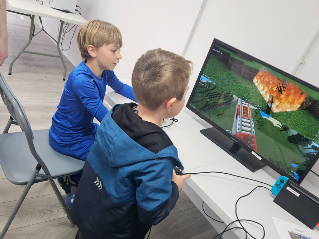 Two boys, one seated and one standing, play a video game on a monitor in a room with several computers. The mood is focused and engaging.