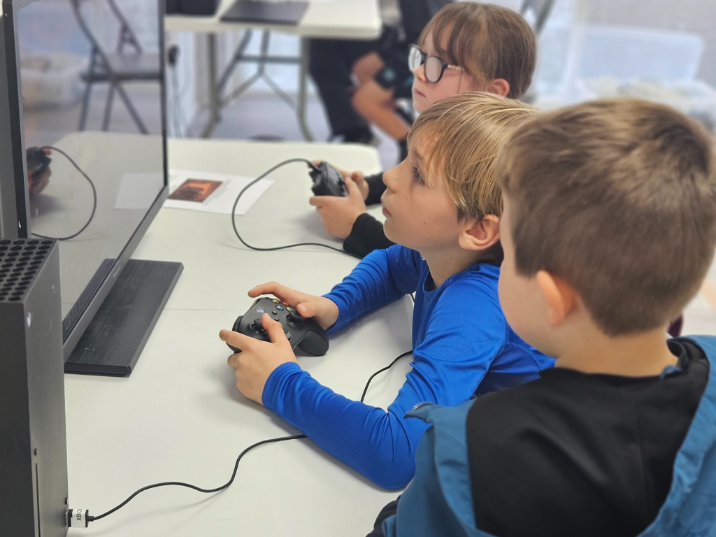 Three children intensely play video games, holding controllers, seated at a table with a large screen. The setting is bright and focused.