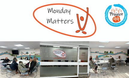 A community gathering at "Monday Matters," showing people seated at tables in a bright room with a mural. Positive and welcoming atmosphere.