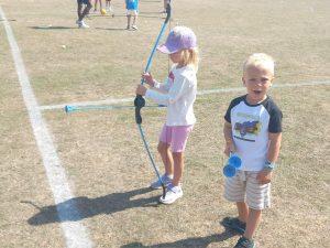 Young girl in a pink cap holds a blue toy bow, standing on a grassy field. A boy in striped shorts stands nearby with a blue toy trumpet.