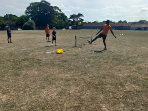 Four people are playing a ball game on a dry grassy field. One person is kicking the ball over a low net. A bucket and rackets lie on the ground.