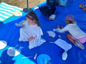 Two young children sitting on a blue tarp, engaged in crafting. They are surrounded by paper plates, glue sticks, and scissors, creating a playful atmosphere.