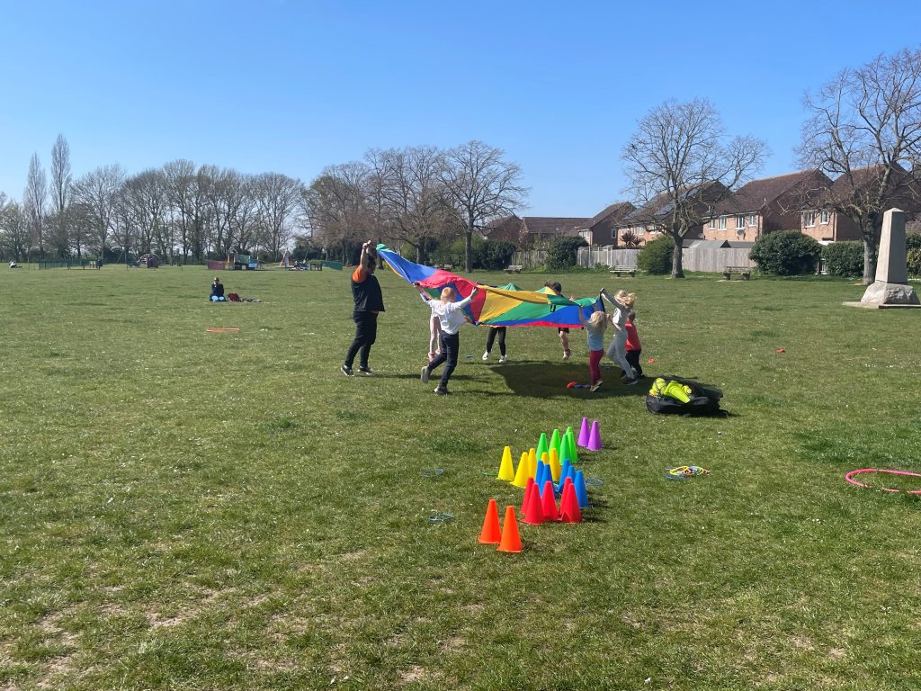 Children play with a colourful parachute in a sunny park. Bright cones are scattered on the grass, and the atmosphere is cheerful and energetic.